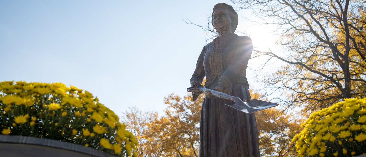 Bronze statue outdoors of a woman in historic clothing, holding a shovel.