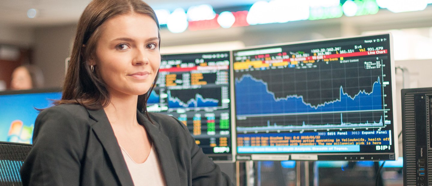 A young woman seated in front of computer screens with colorful charts of data displayed.