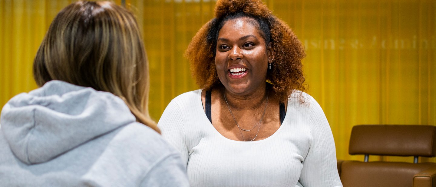 Two women facing each other, talking, in a room with yellow curtains on the walls.