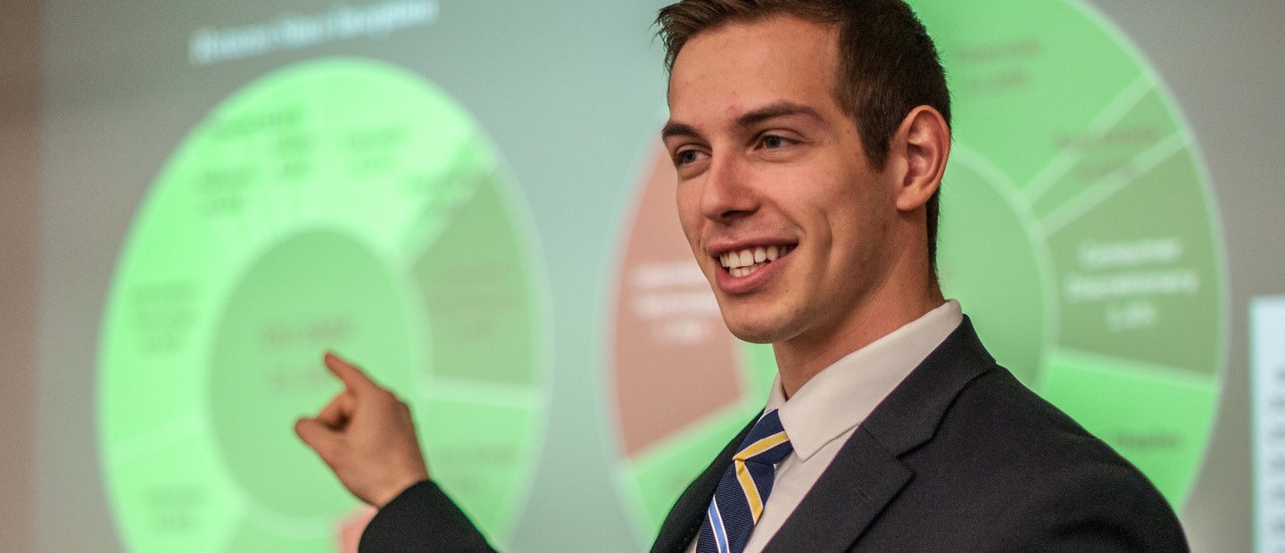 A young man in a suit, pointing to a neon green chart on a screen behind him.