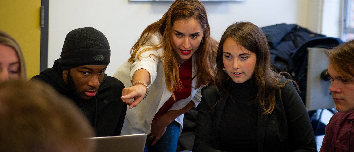 A group of people looking at something on a laptop that one woman is pointing to.