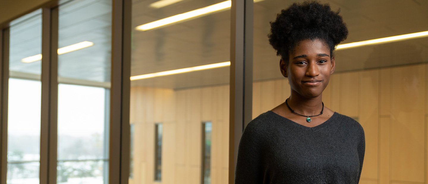 A young woman standing in a room with glass walls, smiling at the camera.