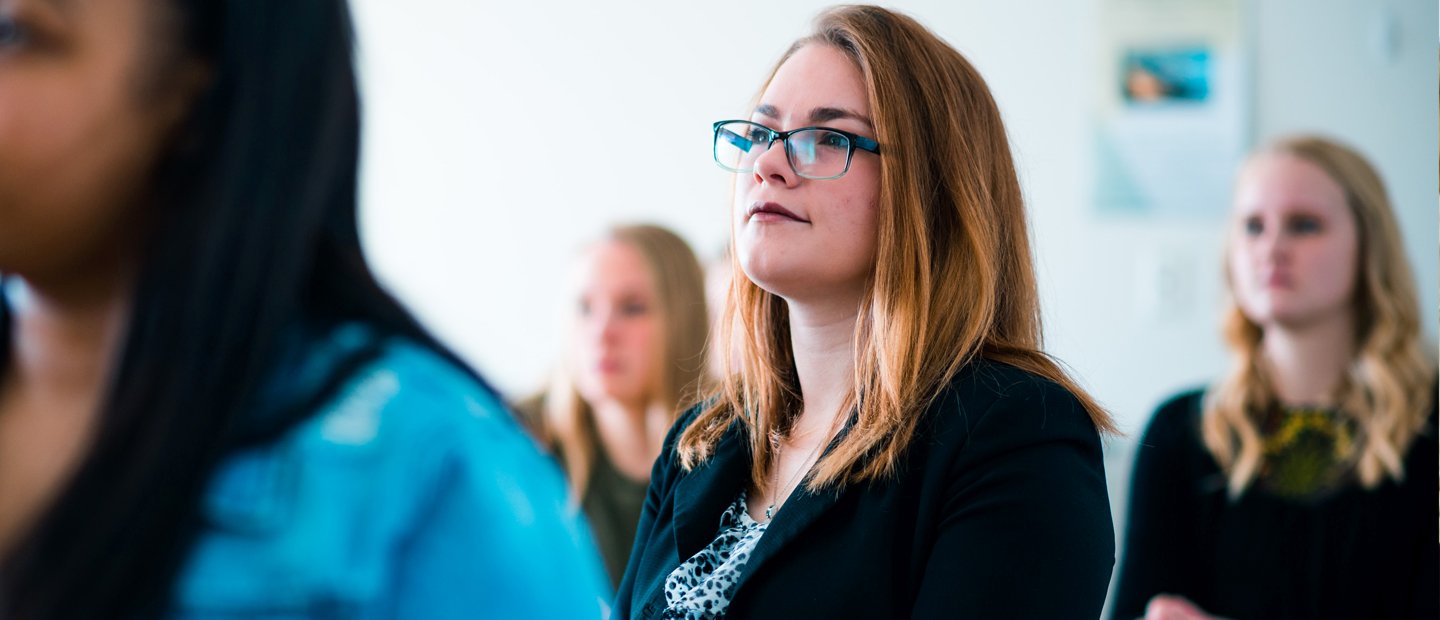 A woman seated at a desk looks towards the front of a classroom.
