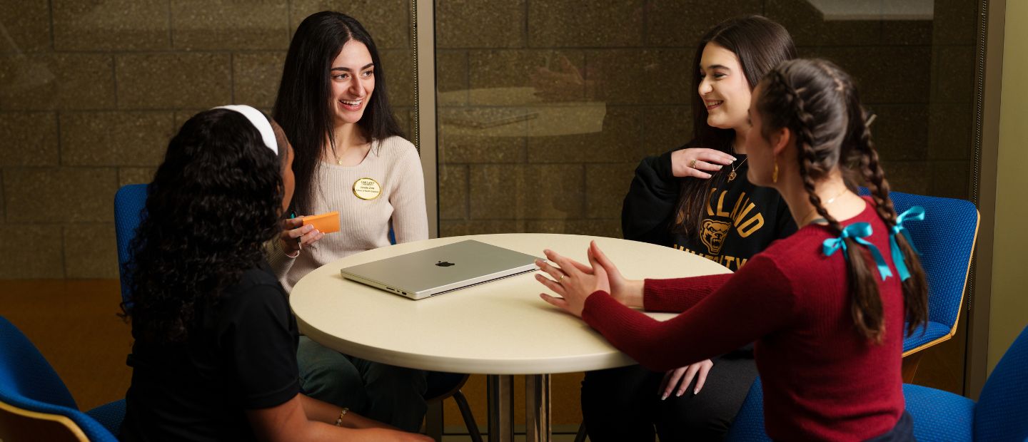 A group of people seated around a table with an open laptop, with others in the background working at computers.