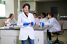 Arwa Hadid in a white lab coat and blue gloves in a Clinical and Diagnostic Sciences lab with other students seated behind her.