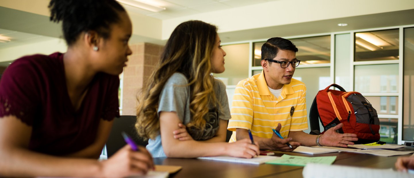 Two students sitting at a table look at a third who is speaking